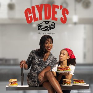 Two women in a lively kitchen—one seated with a knife, the other holding sandwiches