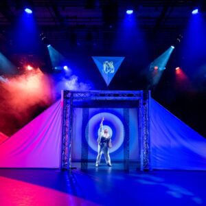 Performer in silvery jumpsuit, centre stage, posed with one arm and head pointed up to blue spotlights