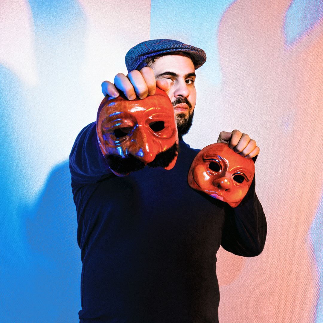 Alaa Shehada, wearing a black shirt and grey newsboy cap, holding two wooden masks.