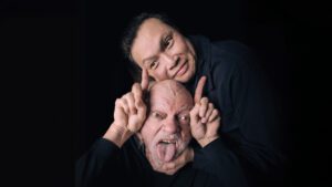 two men in black background, one has his tongue out with hands beside his head and fingers pointed upward, another man rests his cheek on top of the other mans head, looks to his left with a small smile and hand resting on the other persons chin