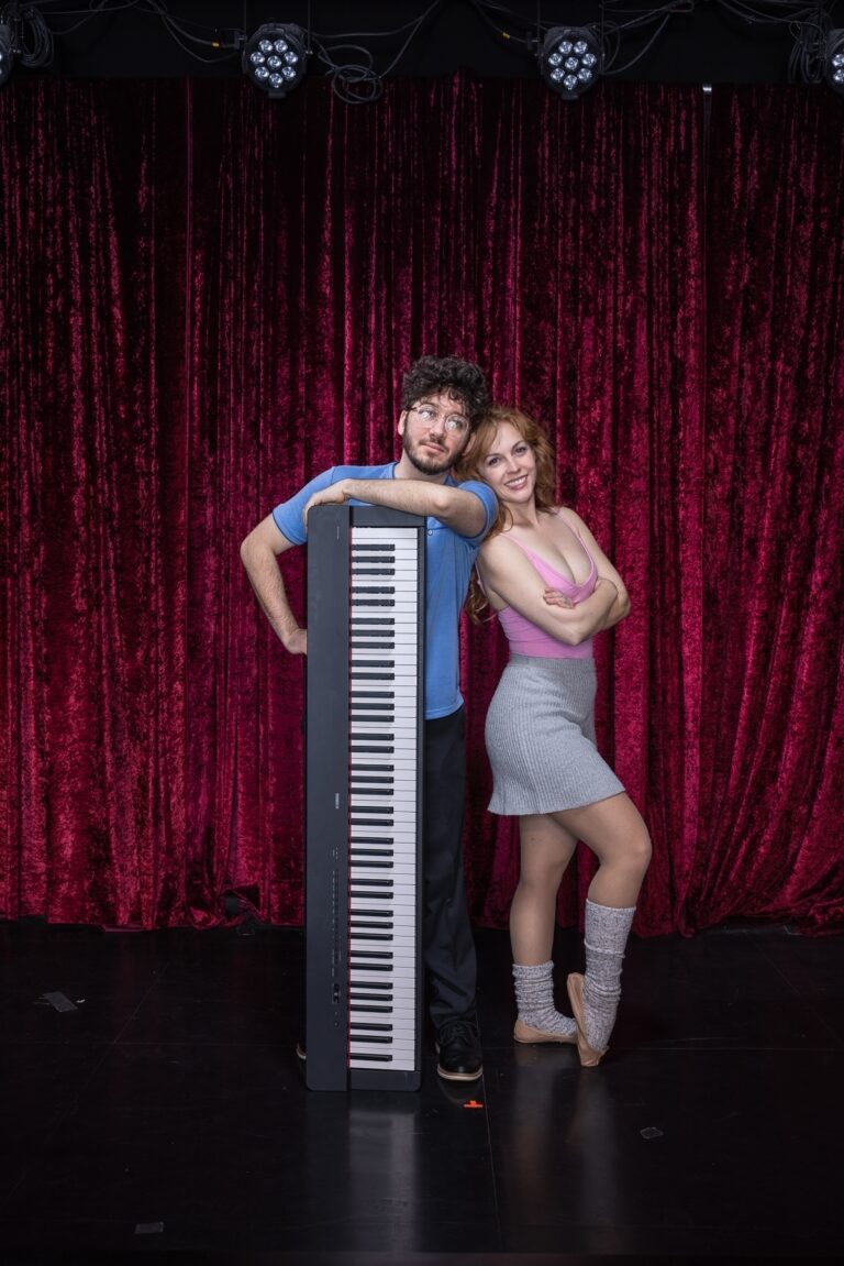 A guy and a gal lean against a piano