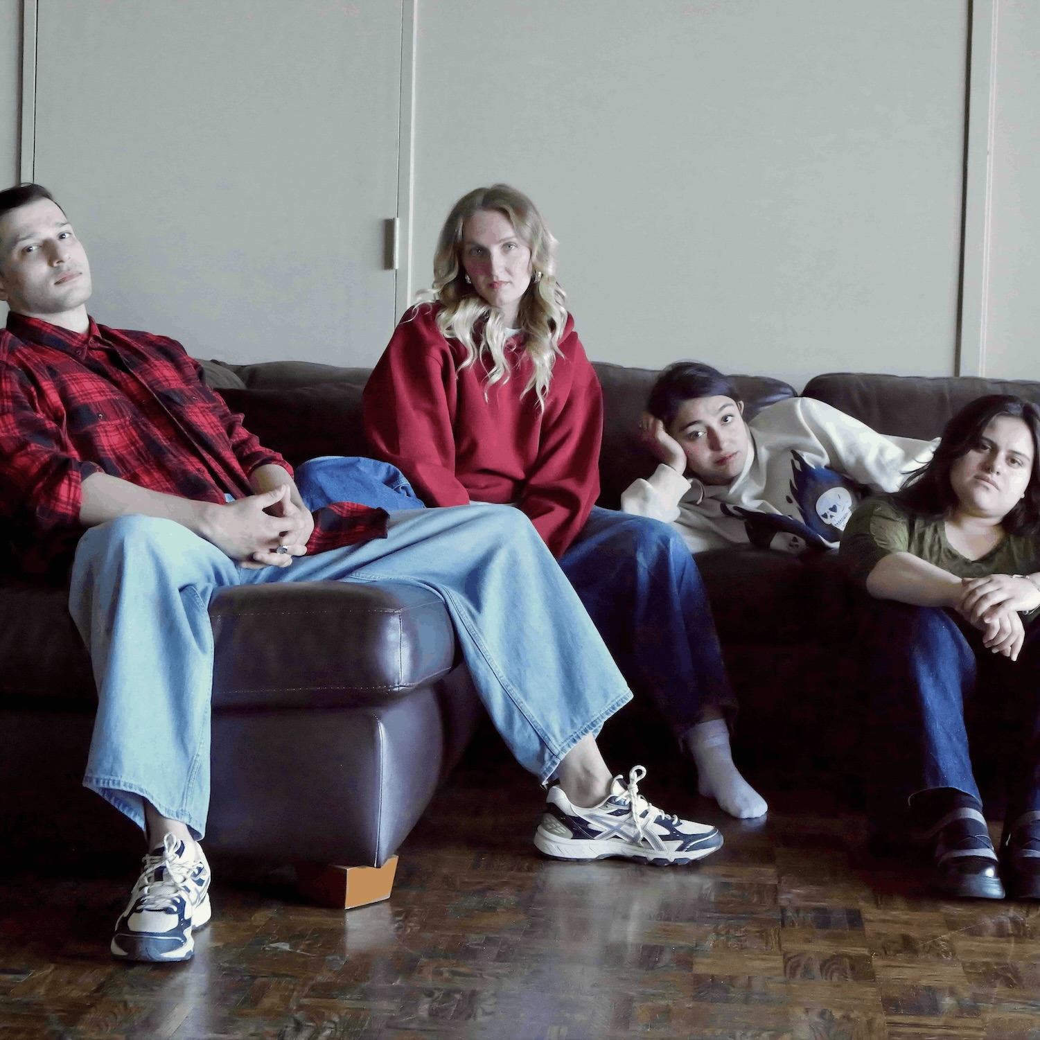 Four people are sitting in a casual, relaxed pose on and around a dark brown leather couch against a plain, neutral-colored wall. 
