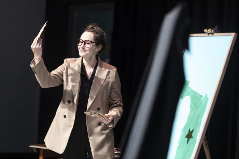White woman with brown hair wearing a tan blazer and red-framed glasses and holding something up in the air 