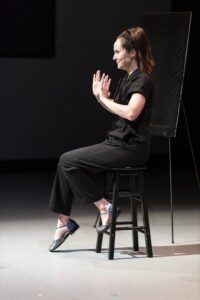 Woman wearing black jumpsuit sitting in a stool with a black theater background