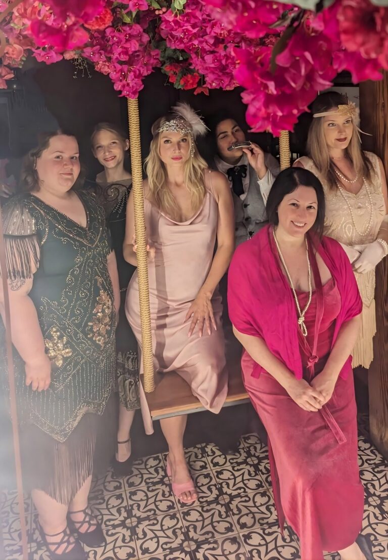 Female people dressed in 1920s gear on and around a wooden swing with pink flowers at top foreground