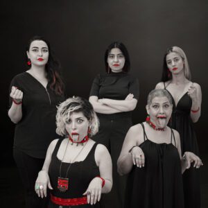 Five women pose against a dark background wearing black clothing with red accents highlighted in selective color. Three stand in the back with composed expressions, while two in the front make playful faces with their tongues out. Red jewelry, lipstick, and accessories contrast with the otherwise black-and-white image.