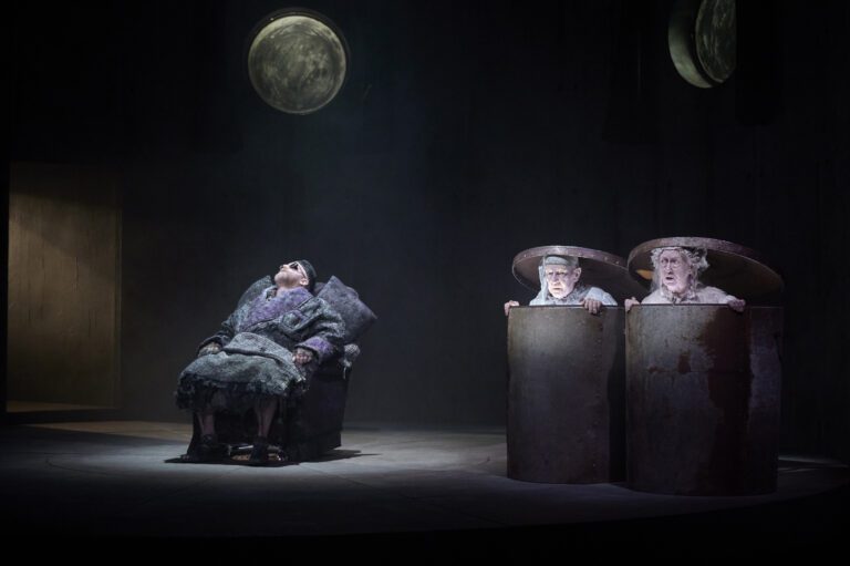 A man is asleep in a chair. Beside him is a man and a woman in sleepwear inside two garbage cans.