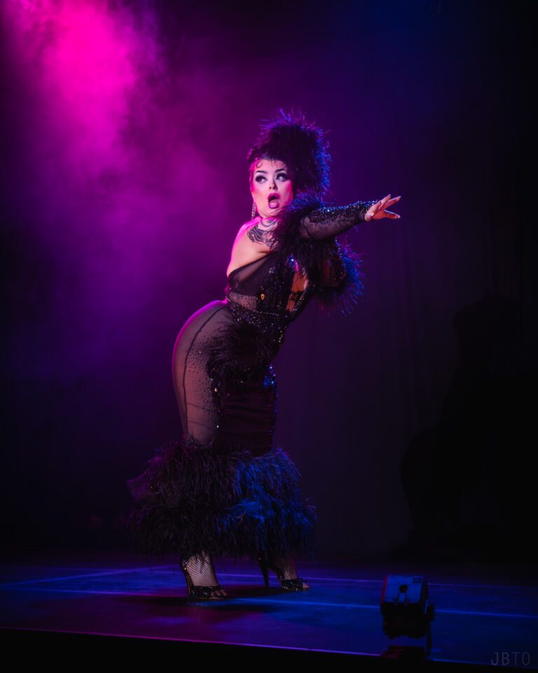 Burlesque performer in a long black gown and black feather boa with a surprised look