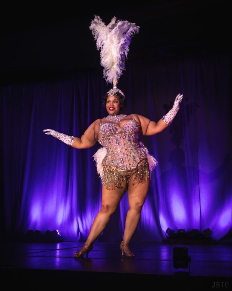Burlesque performer with her arms stretched to the side in a sparkly white costume with a tall feather headpiece
